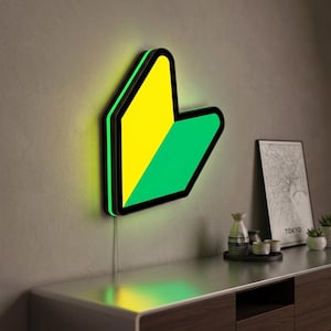 May include: A wall-mounted LED sign shaped like a Japanese beginner driver symbol, featuring a split design with a yellow top-left section and a green bottom-right section, outlined in a thick black border with a glowing light effect.