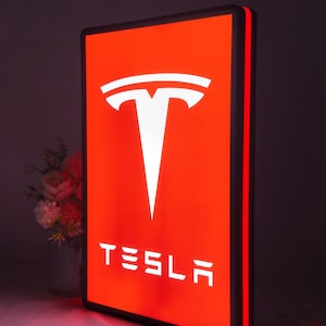 May include: A rectangular, illuminated wall sign featuring a bright red background with the white Tesla logo and the word TESLA printed in white below it. The sign has a black frame and emits a red glow from its side edges, creating a vibrant display.