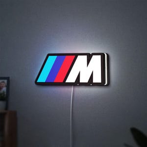 May include: A wall-mounted LED sign featuring the letter M in white, preceded by three slanted stripes in light blue, dark blue, and red. The sign has a black border and emits a soft glow against the wall, with a white power cord extending downward.