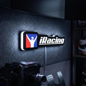 May include: A rectangular wall-mounted LED sign featuring the iRacing logo in white text next to a square icon of a driver with arms raised against a red, white, and blue background. The sign emits a bright white glow from its edges.