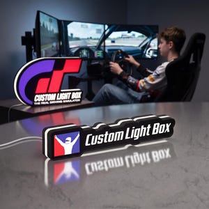 May include: A rectangular LED light box featuring a black frame with white accents. The left side displays a square icon of a white racing driver silhouette against a red and blue background. The text Custom Light Box is printed in white.