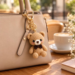 Può includere: Una borsa beige con un portachiavi in metallo dorato che presenta un piccolo orsacchiotto all'uncinetto color marrone chiaro. L'orso ha occhi a perla neri, un naso ricamato marrone scuro, un papillon marrone e guance ricamate rosa.