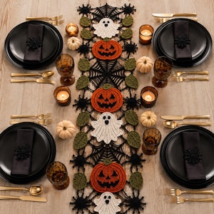 May include: A Halloween-themed crochet table runner featuring alternating white ghosts and orange jack-o'-lanterns connected by black spiderwebs and green leaves, set on a table with black plates, gold cutlery, and small white pumpkins.