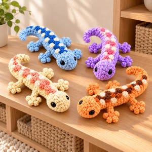 May include: Four crocheted lizard toys with large black eyes. One is blue and white, one is purple and pink, one is cream and peach, and one is orange and brown. Each lizard has a textured, bumpy surface and a long, curved tail with four legs.
