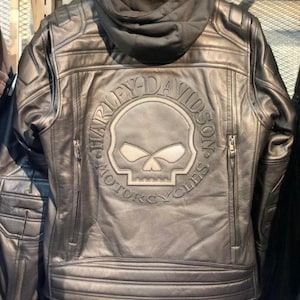 May include: A black leather jacket featuring a hood and a large embossed Harley-Davidson Motorcycles logo on the back. The design includes a central skull graphic, quilted shoulder panels, and two vertical zipped pockets on the lower back.