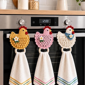 May include: Three crochet chicken towel hangers hanging on an oven handle. Each features a different colored chicken—yellow, pink, and white—with a red comb, a small white flower, and a wooden ring holding a white waffle-weave towel with colored trim.