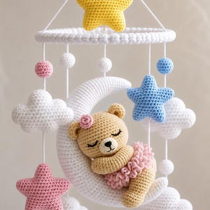 Può includere: Una giostrina per culla all'uncinetto con una mezzaluna bianca centrale e un orsetto beige addormentato che indossa un tutù e un fiore rosa. La giostrina include nuvole bianche sospese, una stella gialla, una blu, una rosa e piccole sfere colorate.