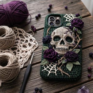 May include: A dark green crocheted phone case featuring a central beige skull motif, surrounded by deep purple crocheted roses, green leaves, and white spiderweb accents. The case is textured with yarn and includes small dark bead embellishments.