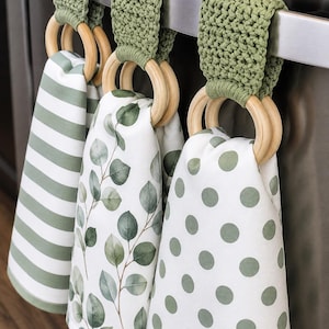 May include: Three hanging kitchen towels featuring sage green patterns: one with horizontal stripes, one with a botanical eucalyptus leaf print, and one with polka dots. Each towel is attached to a light wood ring with a sage green crochet topper.