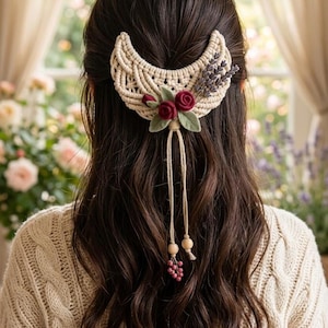 May include: A crescent-shaped macrame hair accessory in off-white cord, featuring two deep red felt roses, green felt leaves, and a sprig of dried lavender. Two thin cords hang down, each finished with a small wooden bead and a cluster of pink berries.