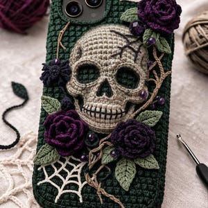 May include: A dark green crocheted phone case featuring a central beige skull, deep purple crocheted roses, green leaves, and white spiderweb accents. The design includes small purple beads and brown vine-like details on a textured yarn surface.