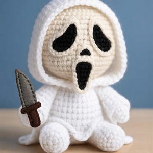 May include: A crocheted doll featuring a white hooded robe and a beige face with black stitched eyes and a black open mouth. The doll holds a small gray felt knife with a brown handle in its right hand, crafted with detailed yarn textures.