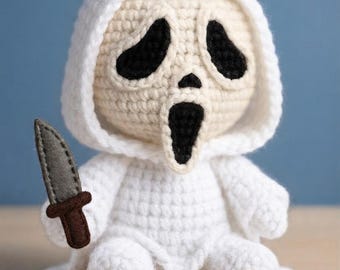 May include: A crocheted doll featuring a white hooded robe and a beige face with black stitched eyes and a black open mouth. The doll holds a small gray felt knife with a brown handle in its right hand, crafted with detailed yarn textures.