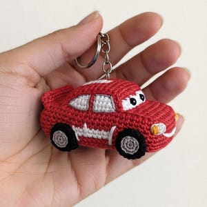 Può includere: Un portachiavi a forma di auto lavorato all'uncinetto in rosso con dettagli dei finestrini bianchi, ruote nere e grigie e un anello in metallo argentato. L'auto ha occhi bianchi con pupille nere, paraurti anteriore bianco e una striscia bianca.
