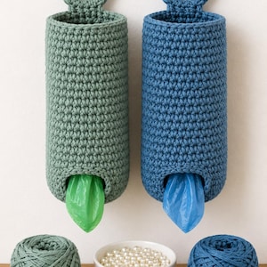 May include: Two cylindrical crochet bag dispensers, one sage green and one blue, each featuring a circular top handle. A green plastic bag protrudes from the sage dispenser, while a blue plastic bag extends from the blue one. Two yarn balls and a bowl of beads sit below.