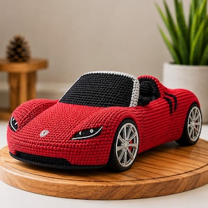 Puede incluir: Un coche de juguete tejido a ganchillo a mano en rojo vibrante con parabrisas negro, interior negro y ruedas plateadas. El coche presenta costuras detalladas, una silueta de coche deportivo de perfil bajo y un pequeño emblema en el capó delantero.