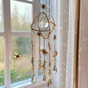 Op de afbeelding: Een goudkleurige metalen mobiel met een centraal bolvormig frame en een gefacetteerde heldere kristallen bol. Meerdere hangende kettingen zijn versierd met kleine gouden zon- en maanbedels, heldere kristallen kralen en langwerpige druppelvormige glasprisma's.