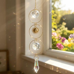 May include: A hanging suncatcher featuring a gold-toned chain with three circular metal rings. The top and bottom rings contain clear faceted crystal spheres, while the middle ring holds a gold sun charm. A clear crystal prism hangs at the base.