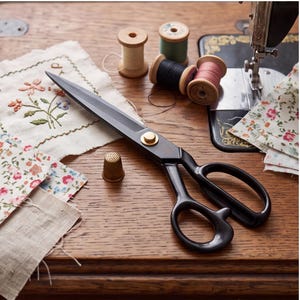 May include: Black sewing scissors with a gold-toned pivot screw, accompanied by a brass thimble, several wooden spools of thread in cream, green, black, and pink, and pieces of fabric with floral embroidery and patterns on a wooden surface.