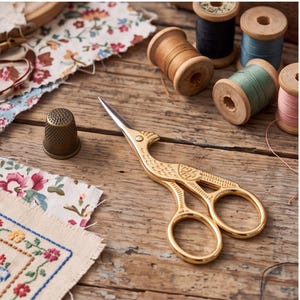 May include: Gold-colored metal scissors shaped like a peacock with intricate engraved feather details on the handles and blades. A metallic thimble and several wooden spools of colored thread are positioned nearby on a rustic wooden surface.