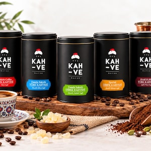 May include: Five black cylindrical coffee tins labelled KAH-VE with a red fez logo. The labels feature different colours: red, blue, green, orange, and maroon. A patterned ceramic cup of coffee, a wooden bowl of mastic, and a wooden scoop of coffee grounds.