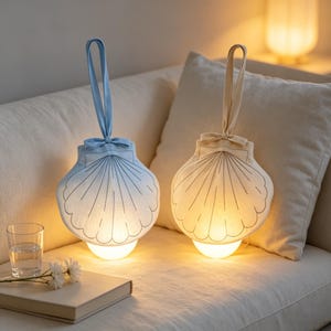 Może przedstawiać: Dwie lampy z tkaniny w kształcie muszli z pętelkami do zawieszania i kokardkami. Jedna lampa jest jasnoniebieska, a druga kremowa, obie z przeszywanymi wzorami muszli i świecącymi podstawami emitującymi ciepłe światło na miękkiej powierzchni.