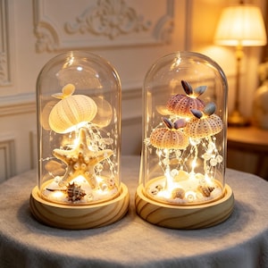 May include: Two glass cloches on round wooden bases, each containing a display of sea urchin shells, a starfish, and small seashells illuminated by thin string lights. The arrangement rests on a white sandy base inside the clear glass domes.