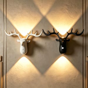 May include: Two wall-mounted deer head lamps, one in matte white and one in matte black, each featuring large antlers. These fixtures emit warm light in upward and downward beams, creating a diamond-shaped glow on the wall surface behind them.