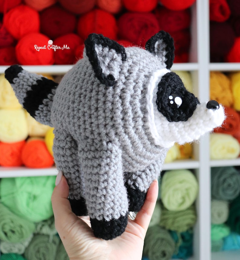 Op de afbeelding: Een grijs en wit gehaakt wasbeer knuffeldier met zwarte oogvlekken, zwarte oortoppen, zwarte pootjes en een gestreepte staart. Het speelgoed heeft een kleine zwarte neus en witte accenten rond het gezicht. De tekst Repeat Crafter Me is zichtbaar.