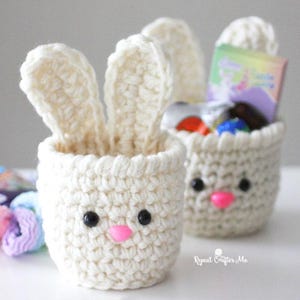 May include: Two handmade crochet bunny baskets in cream color. Each basket has black eyes and a pink nose. One basket has bunny ears and the other has a small gift inside. The baskets are perfect for Easter.