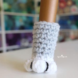May include: A gray and white crocheted cat paw cover. The upper portion is a light gray, while the paw pads are white with black accents. The item is handmade and designed to fit over a doll's foot.