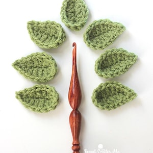 Puede incluir: Un ganchillo de madera con acabado marrón oscuro está rodeado de siete hojas verdes tejidas a ganchillo. Las hojas están dispuestas en semicírculo alrededor del ganchillo. El ganchillo tiene un aspecto liso y pulido.