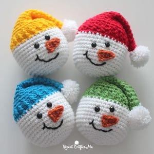 May include: Four crocheted snowman ornaments. Each snowman has a white body, a colored hat (yellow, red, blue, and green), an orange carrot nose, black button eyes, and a stitched smile. Each hat has a white pom-pom.