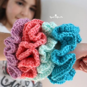 May include: A collection of handmade crochet scrunchies in a variety of colors, including purple, coral, mint green, and turquoise. The scrunchies are made with a textured stitch and are designed to hold hair in place.