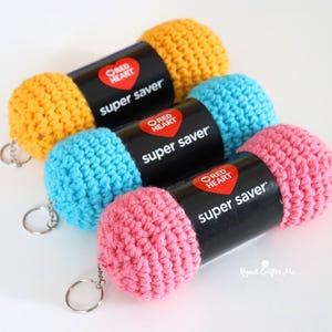 Puede incluir: Tres llaveros de ovillos de lana tejidos a ganchillo en amarillo, azul claro y rosa. Cada uno tiene una etiqueta de papel negro con el logotipo y texto Red Heart Super Saver. Un llavero de metal plateado está unido a cada cilindro.