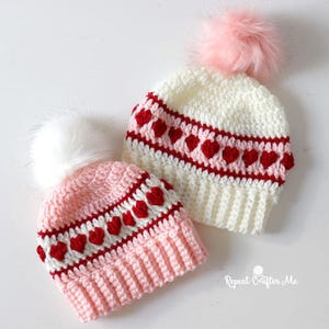 May include: Two handmade crochet beanies. One is pink with a white pom-pom, the other is white with a pink pom-pom. Both have a red heart pattern around the crown. The beanies are likely for babies or children.