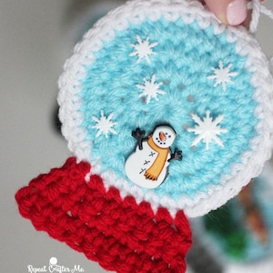 Puede incluir: Adorno navideño de ganchillo hecho a mano que representa un globo de nieve. El globo tiene un centro azul claro con detalles de copos de nieve blancos y un botón de muñeco de nieve. La base es roja y el borde es blanco. El texto "Repeat Crafter Me" es visible.