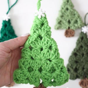 May include: Handmade crocheted Christmas tree decorations in shades of green. Each tree features a brown trunk, a white snowflake detail, and a green loop for hanging. Ideal for festive home decor.