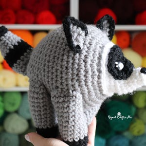 Op de afbeelding: Een gehaakte wasbeer knuffel met een rond grijs lichaam, een gestreepte staart, zwarte oren en een wit gezichtsmasker met zwarte oogvlekken. Het speelgoed heeft een kleine zwarte neus en glanzende zwarte ogen. Tekst: Repeat Crafter Me.