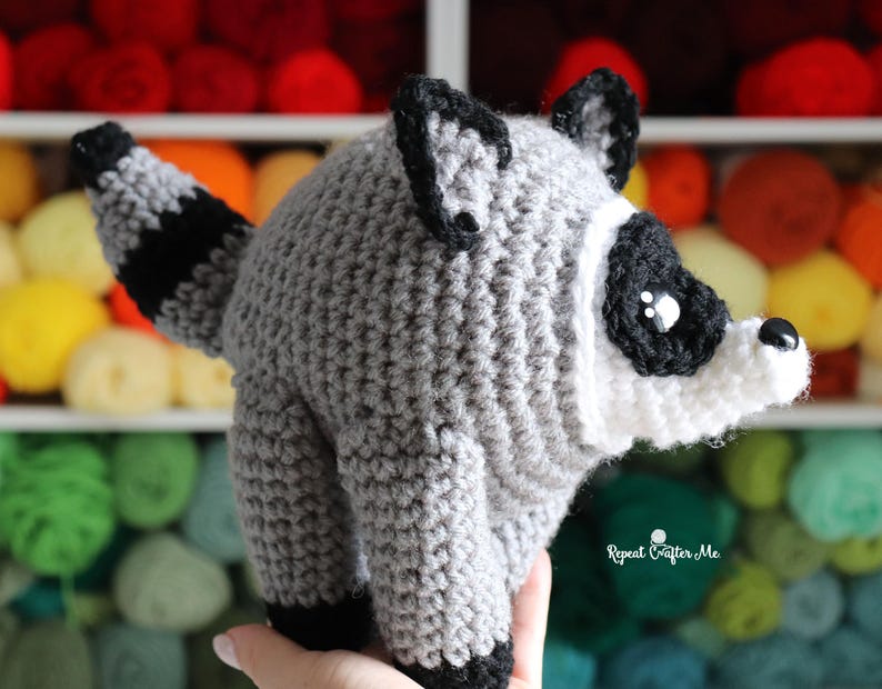 Op de afbeelding: Een gehaakte wasbeer knuffel met een rond grijs lichaam, een gestreepte staart, zwarte oren en een wit gezichtsmasker met zwarte oogvlekken. Het speelgoed heeft een kleine zwarte neus en glanzende zwarte ogen. Tekst: Repeat Crafter Me.