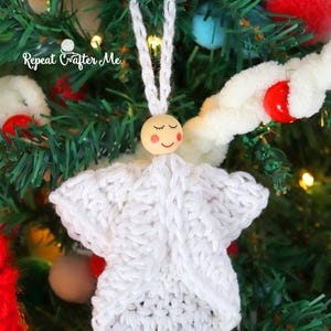 May include: A handmade white crochet angel Christmas ornament. The angel has a wooden bead head with closed eyes and a smiling mouth. The ornament is hanging on a green Christmas tree.