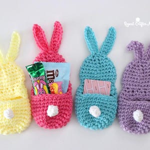 May include: Four handmade crochet bunny-shaped pouches in yellow, pink, blue, and purple. Each pouch has a pocket and bunny ears. The pink pouch contains candy. White pom-poms serve as tails. Ideal for Easter gifts or decorations.