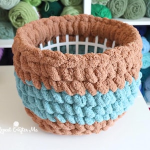 May include: A round storage basket made of thick, chunky yarn in horizontal stripes of brown, light blue, and brown. The basket features a white plastic grid structure visible inside the crochet work. The text Repeat Crafter Me is visible.