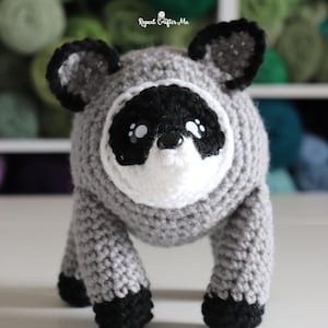 Op de afbeelding: Een gehaakte wasbeer knuffel met een rond grijs lichaam, zwart-witte gezichtsmarkeringen, zwarte oren en zwarte pootjes. Het speelgoed heeft een kleine zwarte neus en witte accenten in de ogen. De tekst Repeat Crafter Me is zichtbaar.
