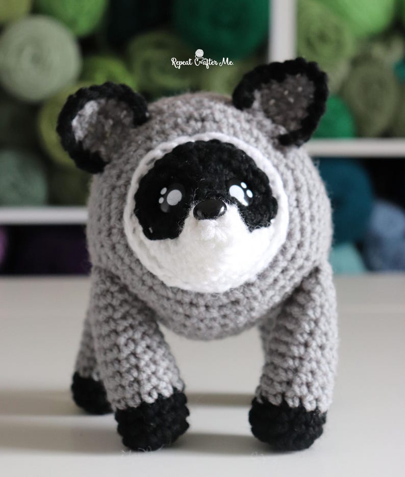 Op de afbeelding: Een gehaakte wasbeer knuffel met een rond grijs lichaam, zwart-witte gezichtsmarkeringen, zwarte oren en zwarte pootjes. Het speelgoed heeft een kleine zwarte neus en witte accenten in de ogen. De tekst Repeat Crafter Me is zichtbaar.