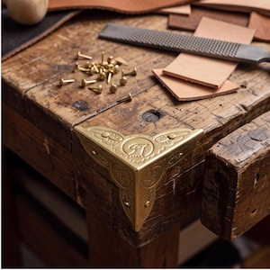 May include: A gold-toned metal corner protector featuring an embossed bird motif and decorative leaf patterns. The hardware is designed for attachment to a corner edge, secured by small brass nails scattered nearby on a wooden workbench surface.