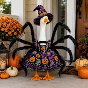 May include: A white goose figurine wearing a purple and black witch hat and a matching dress, both featuring orange pumpkin and white spiderweb patterns. The goose has eight long, black, fuzzy spider legs extending from its back.