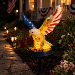 May include: A solar-powered garden stake featuring a bald eagle with wings spread wide. The eagle's left wing is blue with white stars, and the right wing features red and white stripes. The body is illuminated with a warm yellow glow.