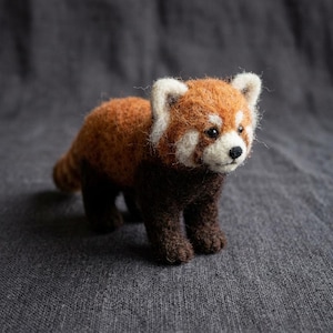 May include: A needle-felted red panda figurine featuring a textured rust-orange back, dark brown legs, and a white face with black eyes and a small black nose. The small animal sculpture has rounded ears and a soft, fuzzy appearance throughout.