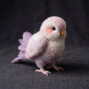 May include: A needle-felted bird figurine with soft, fuzzy light pink and white wool fibres. The bird features a small orange beak, dark round eyes, and a textured, light purple fan-shaped tail. It stands on two small, light-coloured feet.